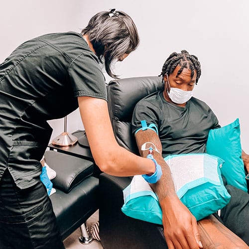 A person wearing a mask sits in a chair with their arm resting on a pillow while a healthcare worker prepares a vitamin infusion or to draw blood. Both individuals are dressed in black clothing.