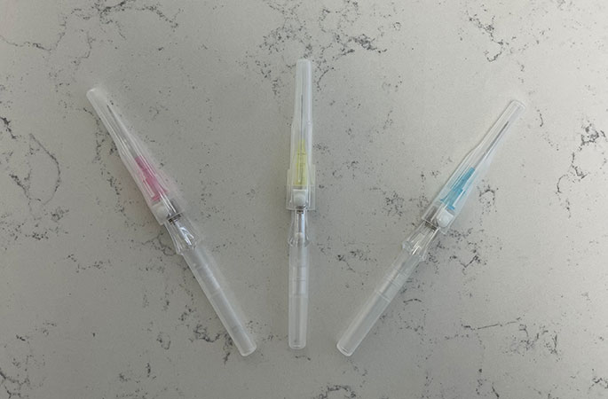 Three capped syringes with colored bands—pink, yellow, and blue—are arranged in a V shape on a light gray, marbled surface, suggesting preparation for a vitamin infusion.
